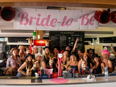Un grupo grande de mujeres vestidas de vaqueras celebrando una despedida de soltera detrás de la barra de un bar con un cartel de "Bride-to-Be" durante un taller de coctelería en Albufeira.