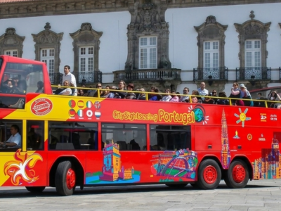 Autobús turístico rojo de dos pisos recorriendo una plaza histórica de Oporto frente a un edificio monumental