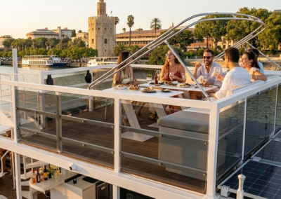 : Grupo de amigos disfrutando de una cena privada en la cubierta superior de un barco solar en Sevilla con la Torre del Oro al fondo.