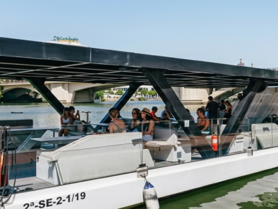 Barco turístico privado atracado en el muelle del río Guadalquivir en Sevilla con pasajeros a bordo.