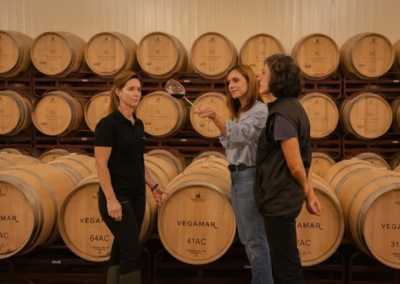 Tres mujeres catan vino tinto rodeadas de barricas de roble en la bodega Vegamar durante una visita guiada en Valencia.