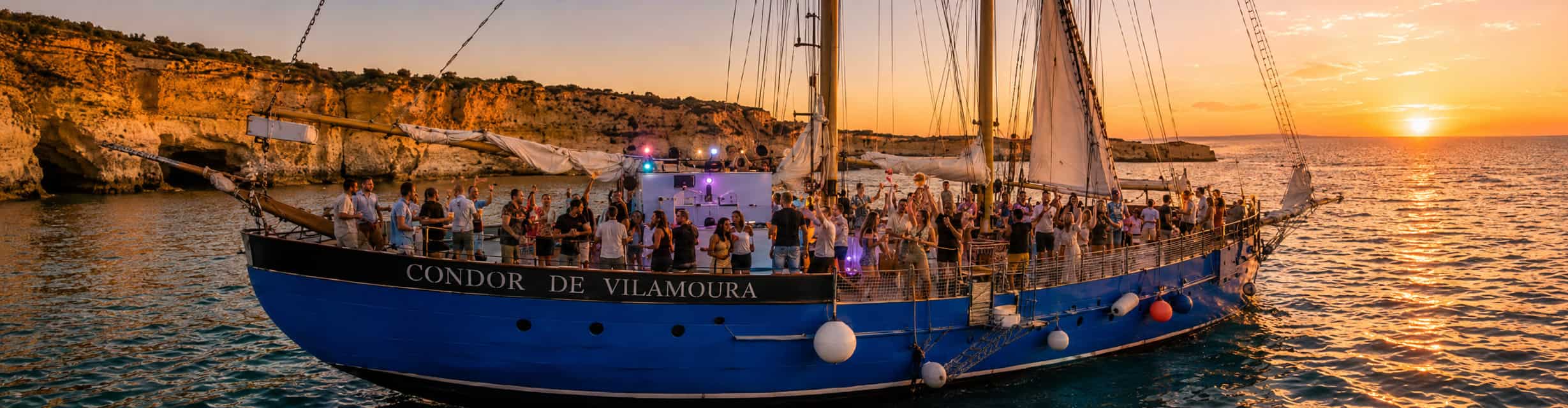 Barco de fiesta al atardecer saliendo de la Marina de Vilamoura con grupos celebrando a bordo y los acantilados dorados del Algarve de fondo · TiToTravel
