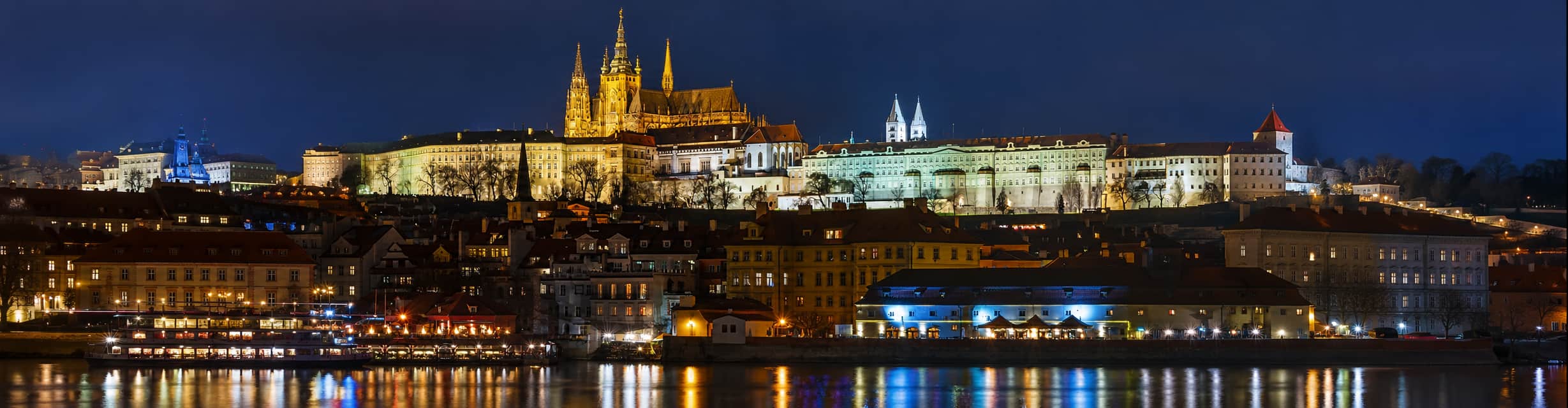 Crucero de cena en barco de cristal navegando por el río Moldava de noche con el Puente de Carlos iluminado de fondo · TiToTravel